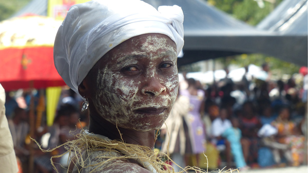 A celebrant with white paste on her face and body, wearing a collar of straw. 